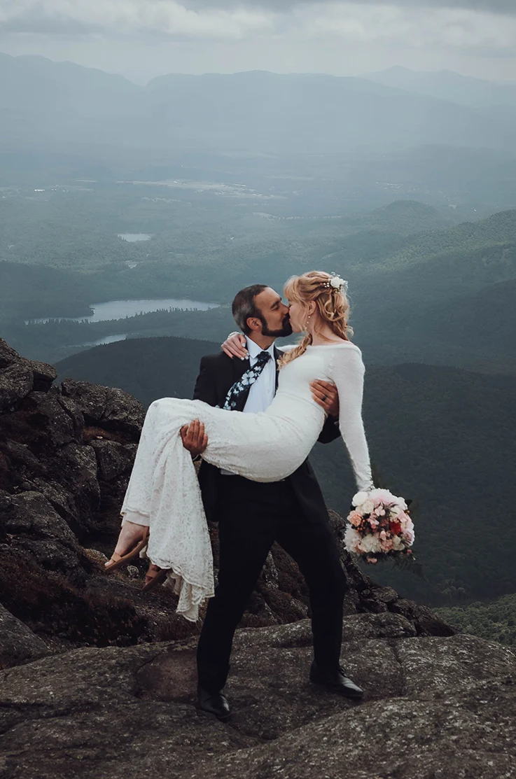 Groom picks up his bride in a fit and flare lace gown with long sleeves, and is holding a bouquet on a rocky mountaintop.