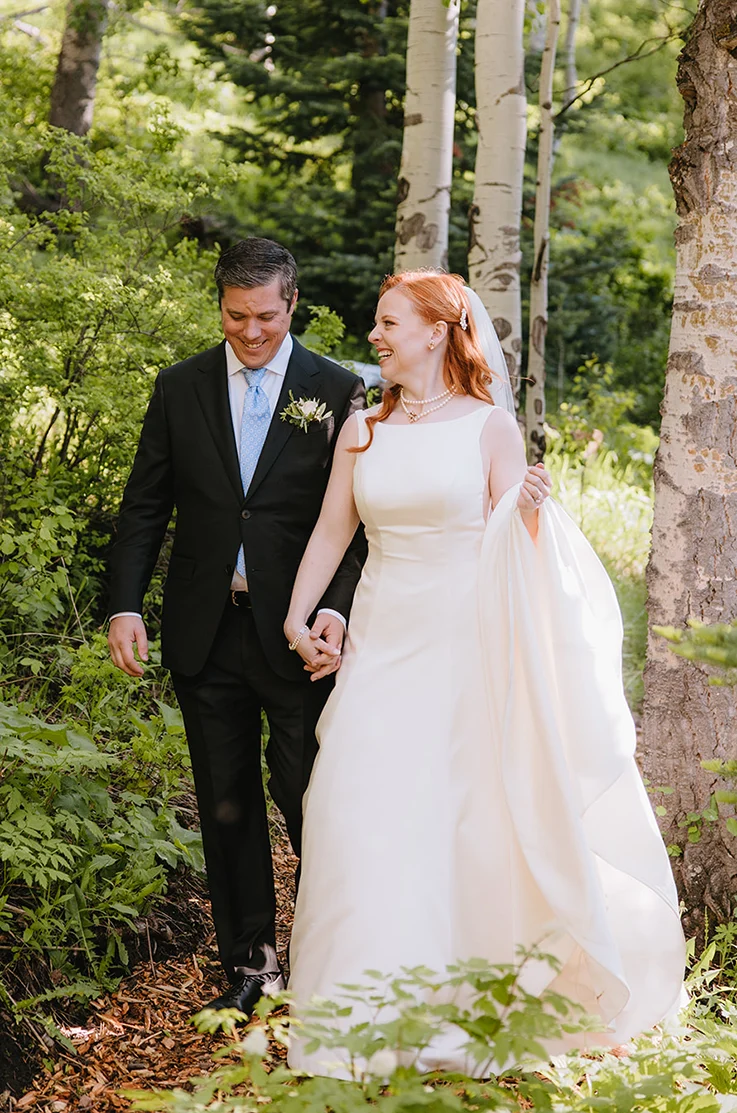 Bride in a satin A-line gown with a bateau neckline and a veil, walks hand in hand with her groom on a forest path.