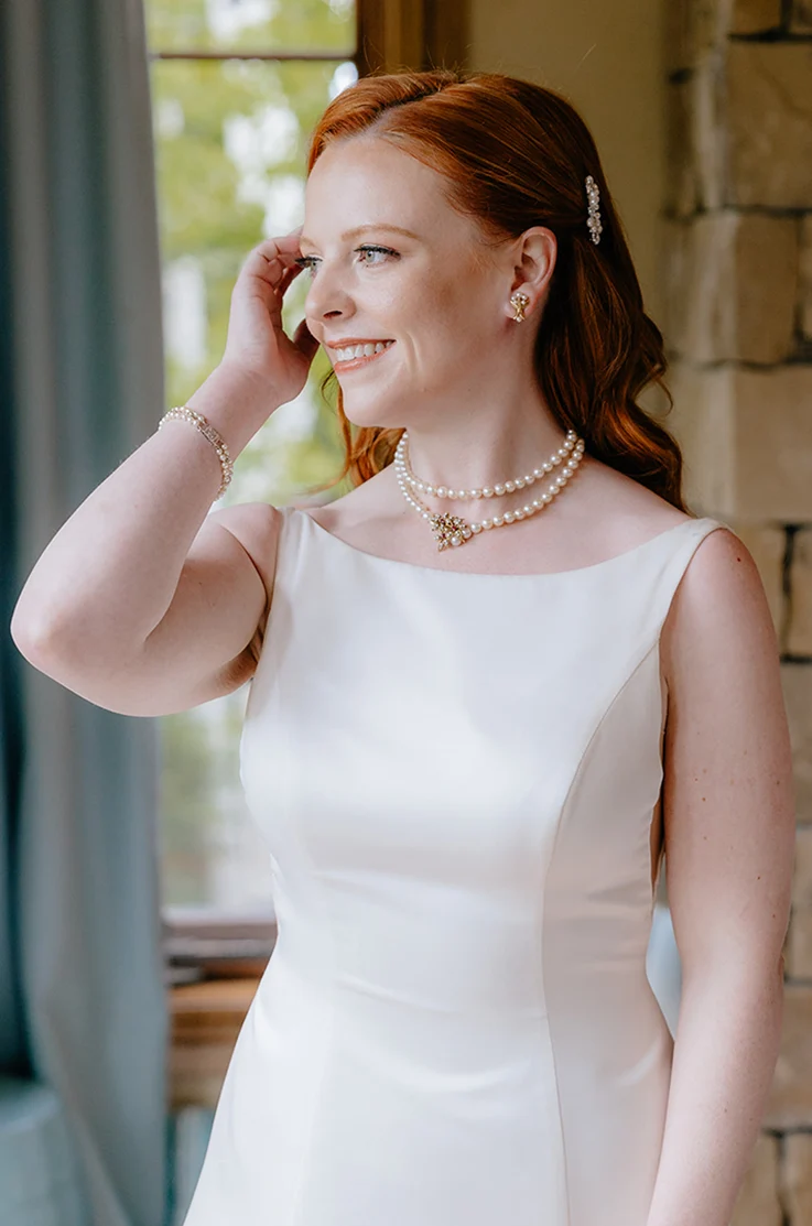 Close-up of bride in a sleeveless satin wedding dress with a bateau neckline, poses in a bridal suite indoors.