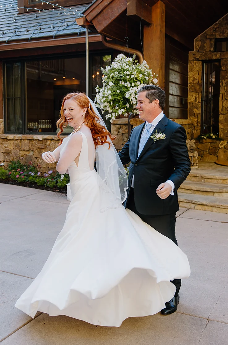 Bride spins in a satin A-line gown with tulle side inserts, a low open back and a veil beside her groom in front of the venue.