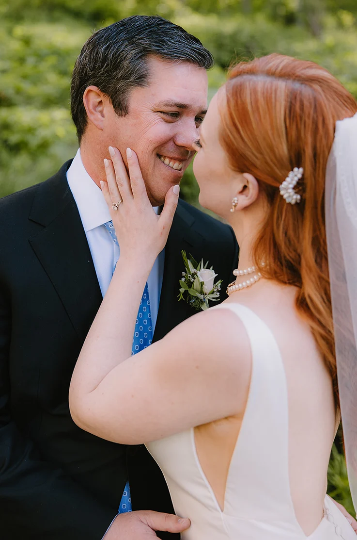 Close-up of bride facing her groom in a low back satin gown with side cutouts and a veil, and they embrace one other outdoors.