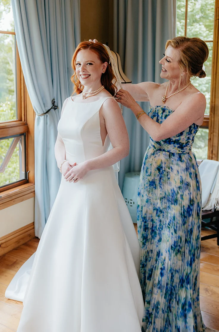 Bride's mother adjusts her veil in a bateau neck satin gown with an A-line skirt by open windows with drawn blue curtains.