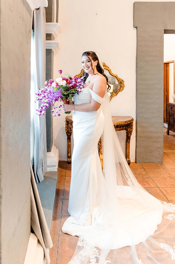 Bride in a crepe draped bodice wedding dress with off-shoulder sleeves and a veil, holds a bouquet in front of a window.