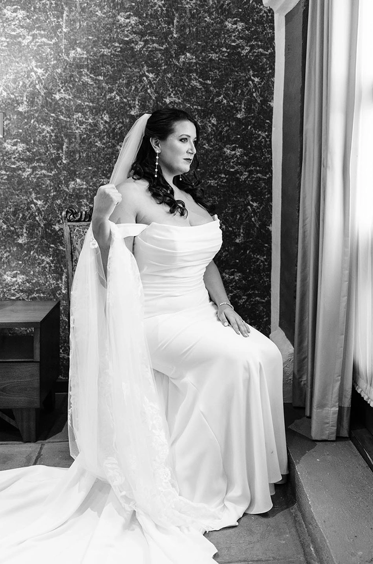 Bride sits on a chair in a draped crepe gown with a scoop neck, sleeves, a veil and fit and flare skirt in black and white. 
