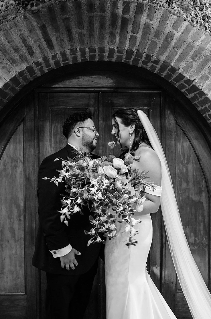 Bride in an off-shoulder fit and flare gown with draped sleeves and a veil, stands with groom in front of venue doors.