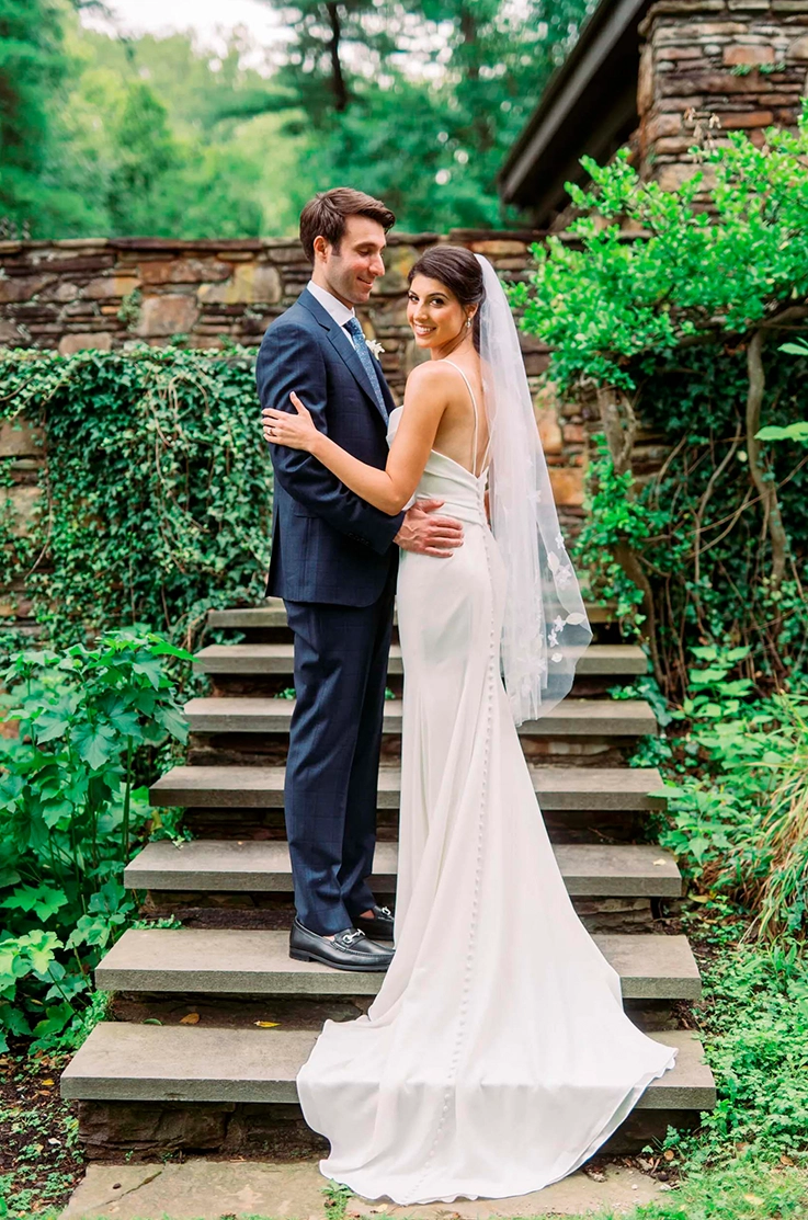 Groom embraces his bride in a spaghetti strap crepe gown with a low buttoned back and a veil, on stone steps.