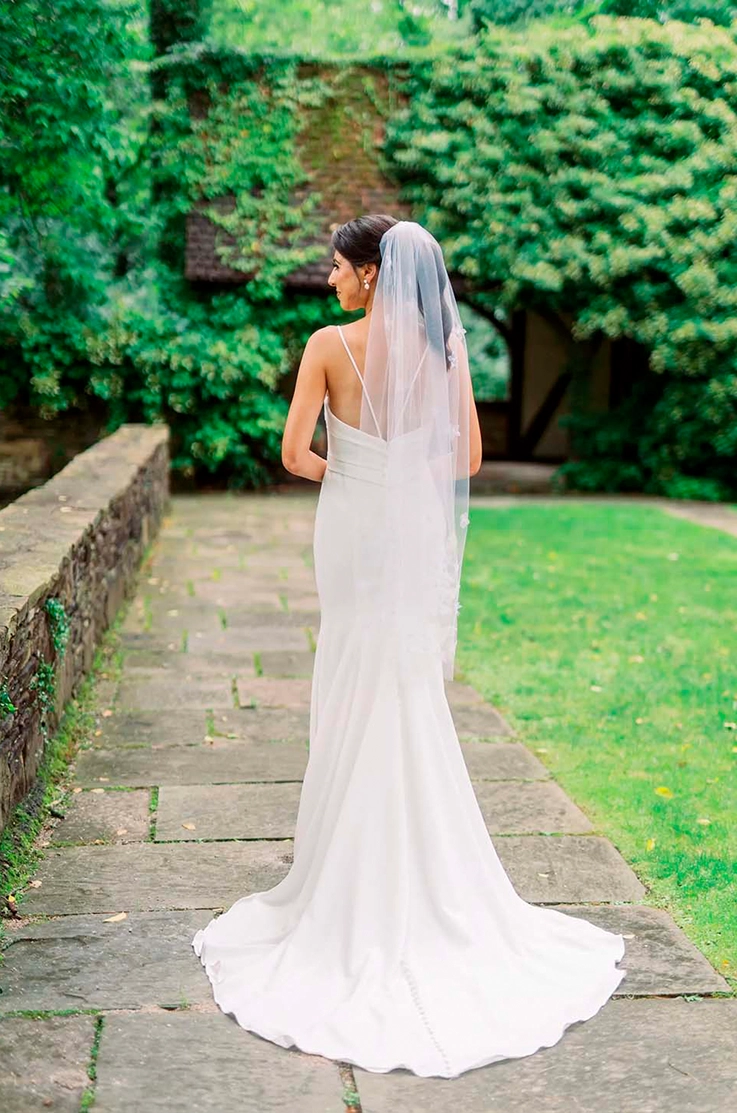 Back of bride in a fit and flare crepe gown with a pleated bodice and veil, is standing on a garden path.