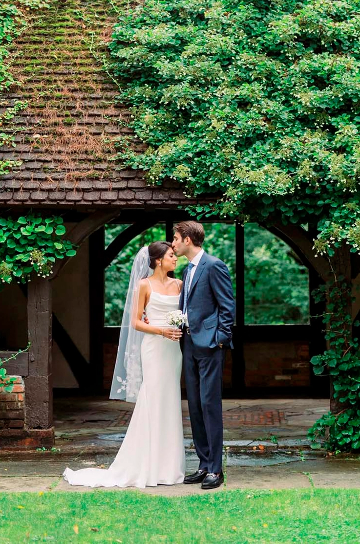 Bride poses in a crepe fit and flare wedding dress with a pleated bodice and a veil, next to groom at an ivy-covered cottage.