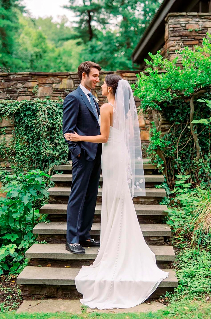 Bride in a crepe fitted gown with a low back and buttoned train with a veil, faces her groom on stone garden steps. 