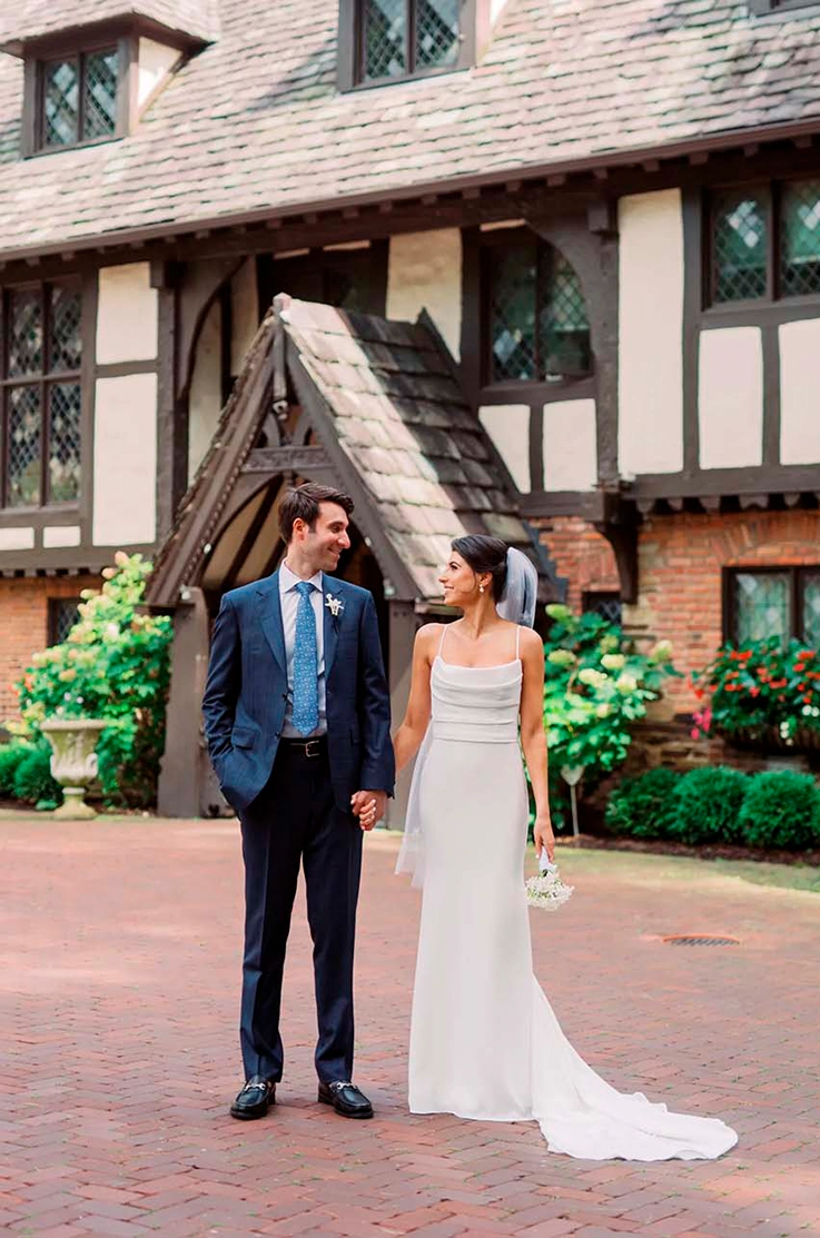 Bride in a crepe pleated bodice with a scoop neck and fit and flare skirt, smiles at her groom outside the venue.