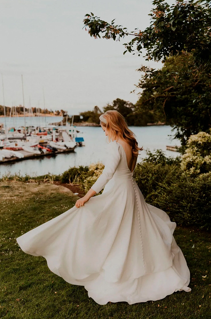Bride twirls in a crepe long cap sleeve gown with buttons and a V-neckline at a marina as the sun sets.