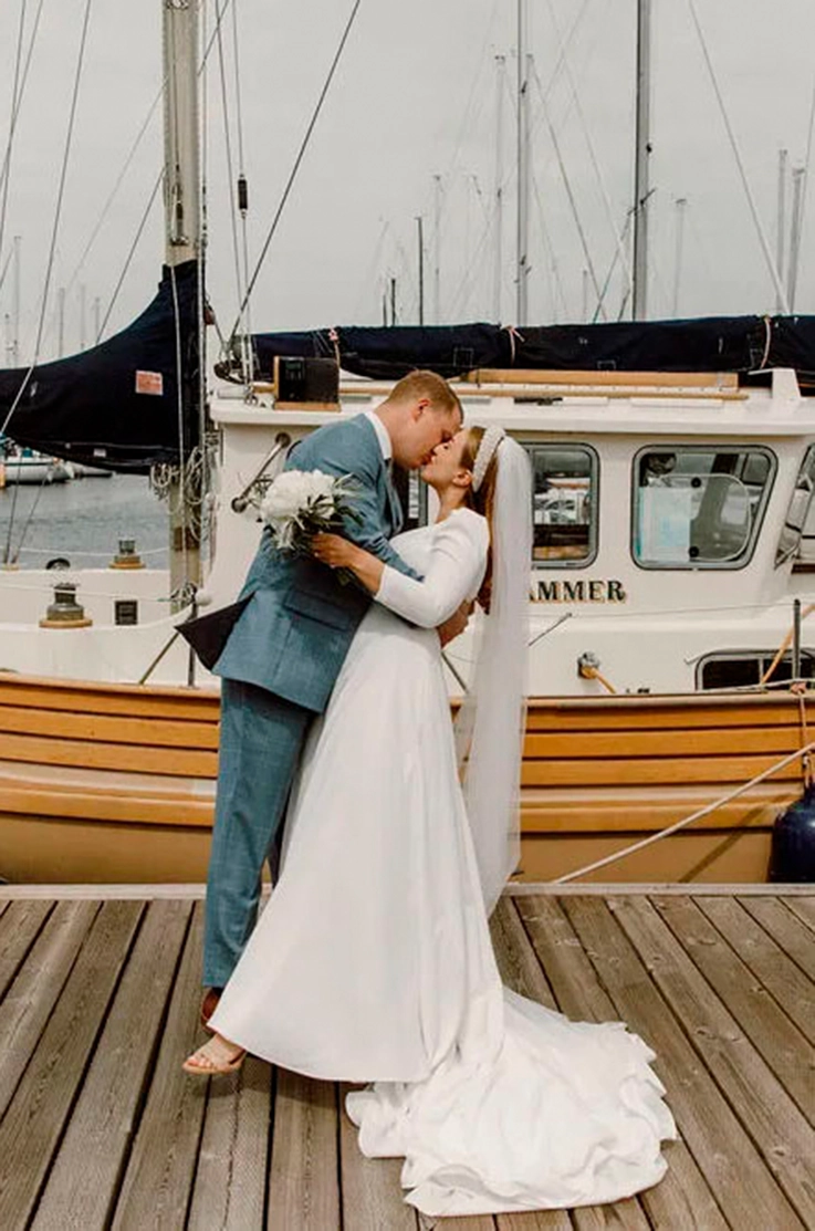 Bride in a crepe sleeved wedding dress with an A-line skirt and veil, kisses groom in a blue suit on a marina dock.
