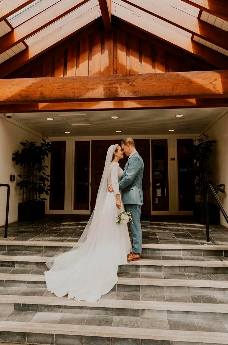 Groom embraces bride in a crepe gown with cap sleeves, a flowing train and veil, as they stand on steps outside the venue.