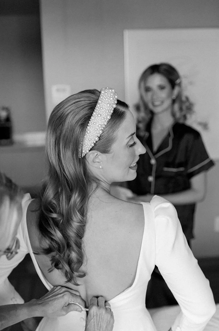 Close-up of bride getting ready in an open back crepe gown with puff sleeves and a pearl headband, indoors in black and white.