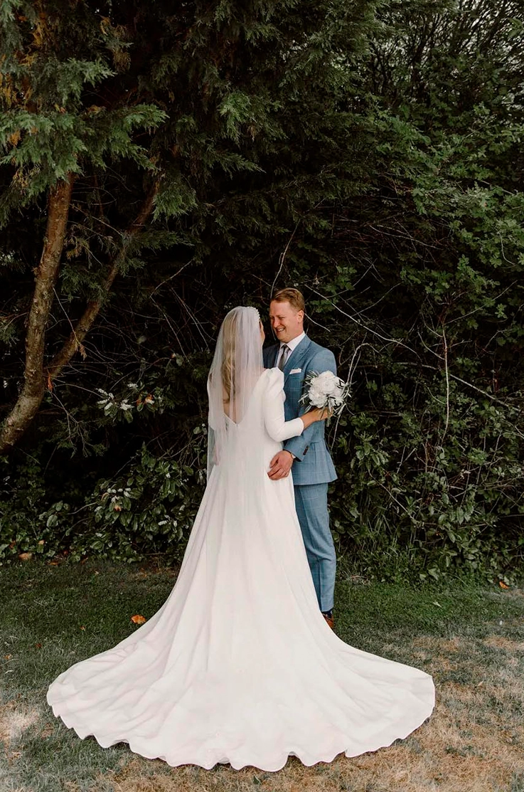 Bride in a deep-V back crepe gown with sleeves and a veil, faces groom in a blue suit outdoors in front of trees. 