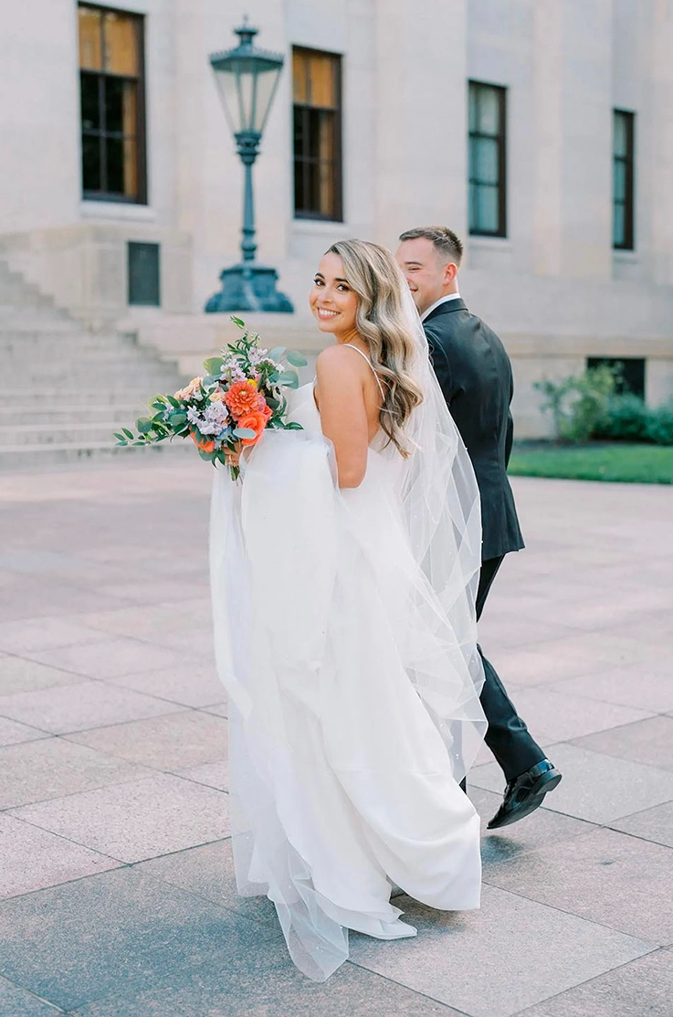Bride holds her train and a bouquet, in a crepe spaghetti strap gown with a veil, and walks beside groom towards the venue.