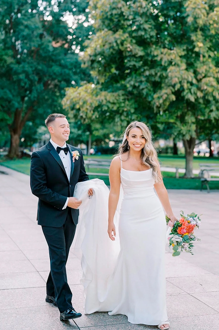 Groom walks alongside bride and holds her train in a scoop neck crepe wedding dress, holding a bouquet on an outdoor path.