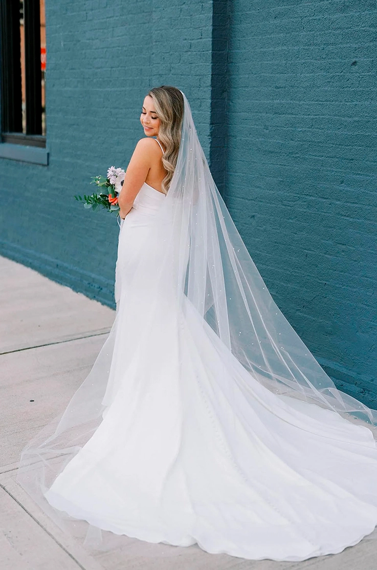 Bride poses and looks past her shoulder in a low back crepe gown with a veil, and is holding a bouquet by a blue brick wall.