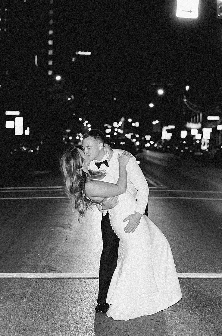 Groom dips his bride in a crepe fit and flare gown with button detail and a scoop neck, on a city street in black and white. 