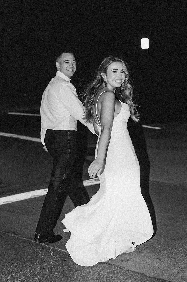 Bride in a pleated crepe bodice gown with a fit and flare skirt and a low back, walks next to groom in black and white.