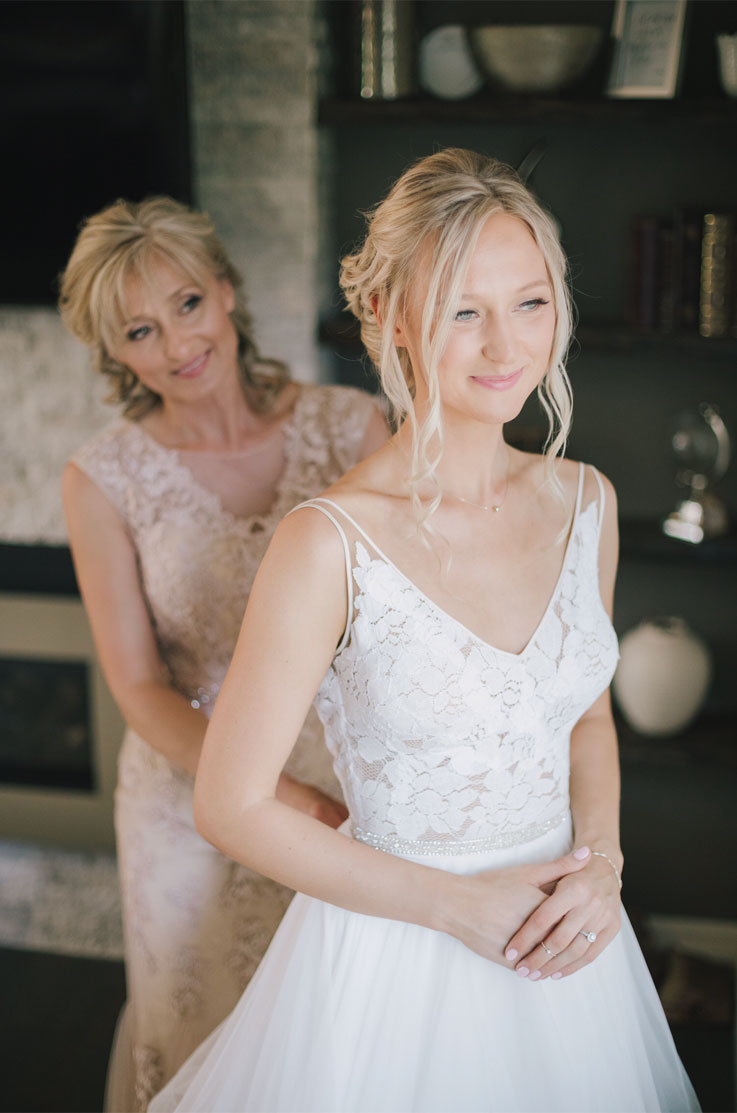 A woman in a lace dress helps zip up the dress of a smiling bride in a lace and tulle wedding dress, while they stand indoors. 