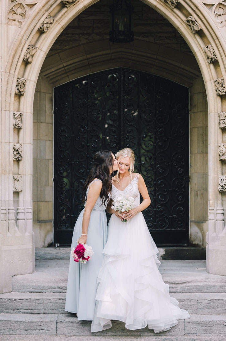 A woman in a Chiffon dress holding a bouquet kisses a bride in a lace and tulle wedding dress on her cheek, as they stand at the top of a stone staircase in front of a building with iron gates.