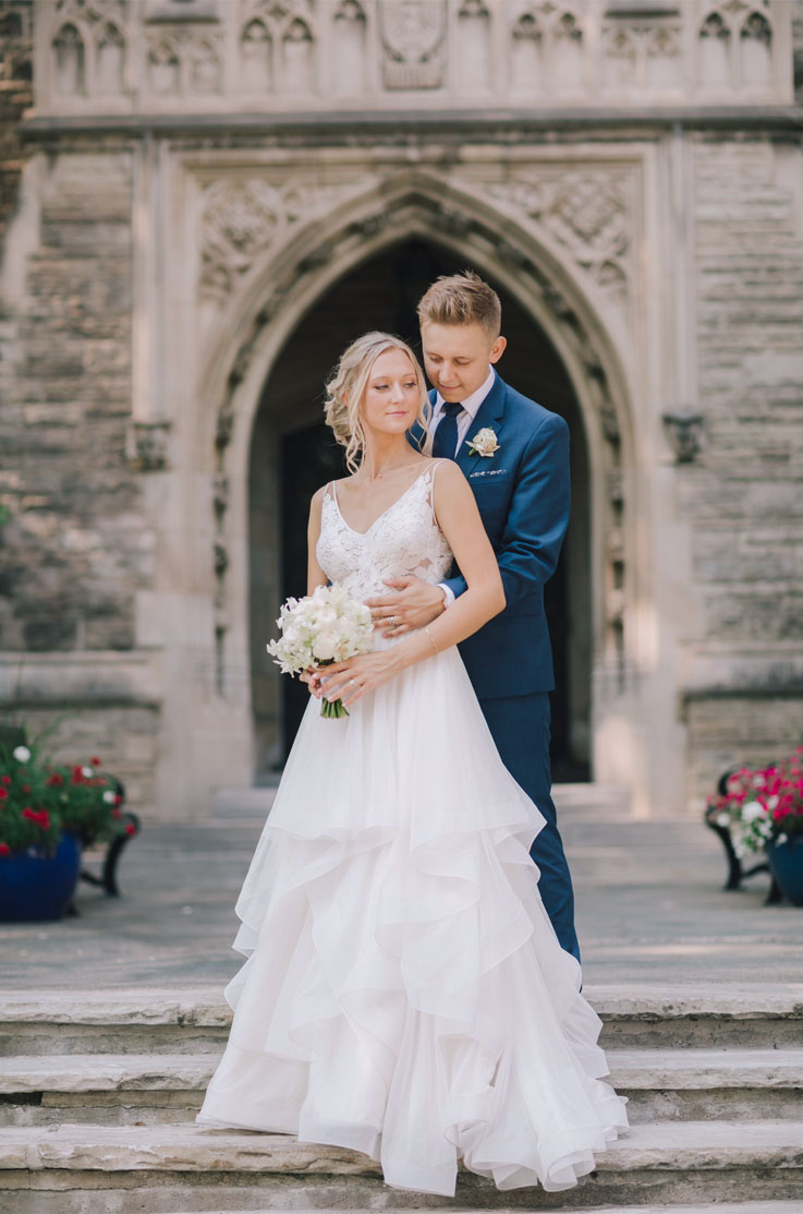 A groom in a suit holds the waist of a bride in a lace and tulle wedding dress, as they stand outside at the top of a staircase in front of a stone building.