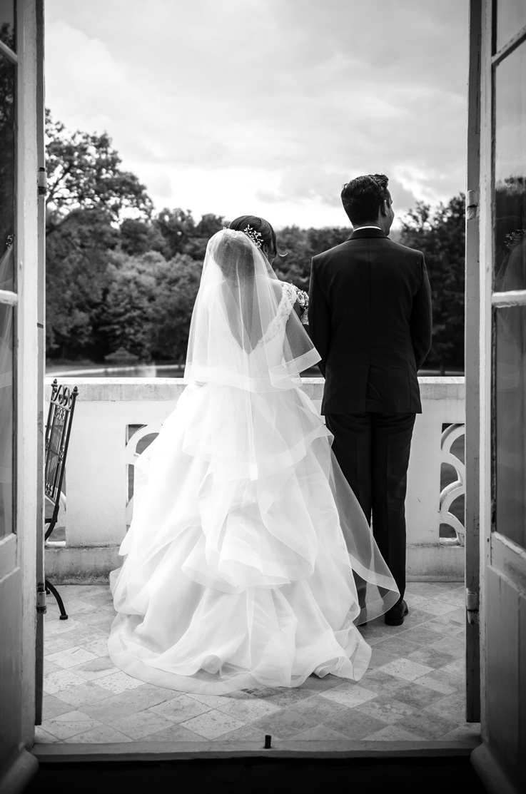 A bride in a tiered ball gown wedding dress and veil, and a groom in a suit, stand on a balcony and look out at the view, with trees in the distance.