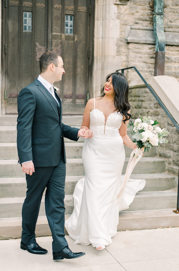 A bride in a plunging neckline wedding dress, holding a bouquet, holds the hand of a groom in a suit as they walk down a small staircase outside of a stone building.