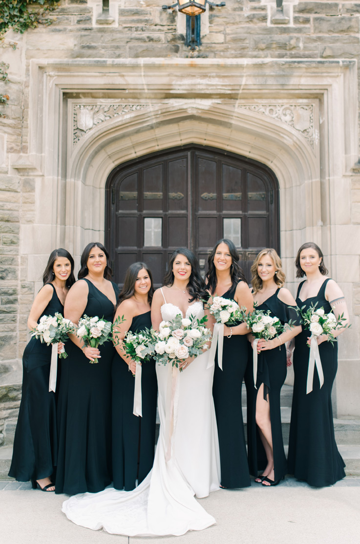 A bride in a fitted wedding dress stands between six bridesmaids, all of them smiling and holding bouquets. They are standing in front of a stone building with wooden double doors.