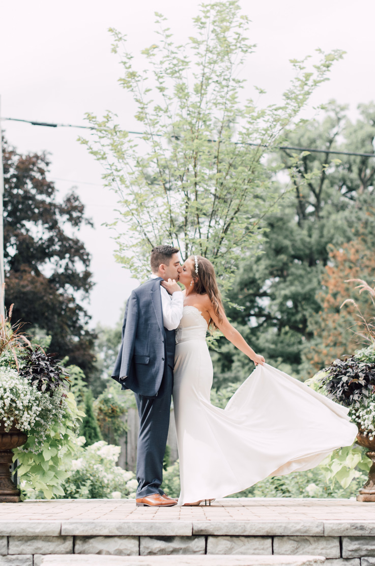 A bride in a strapless wedding dress and a groom in a suit, holding his jacket over his shoulder, kiss on a stone patio outdoors, with trees in the background.