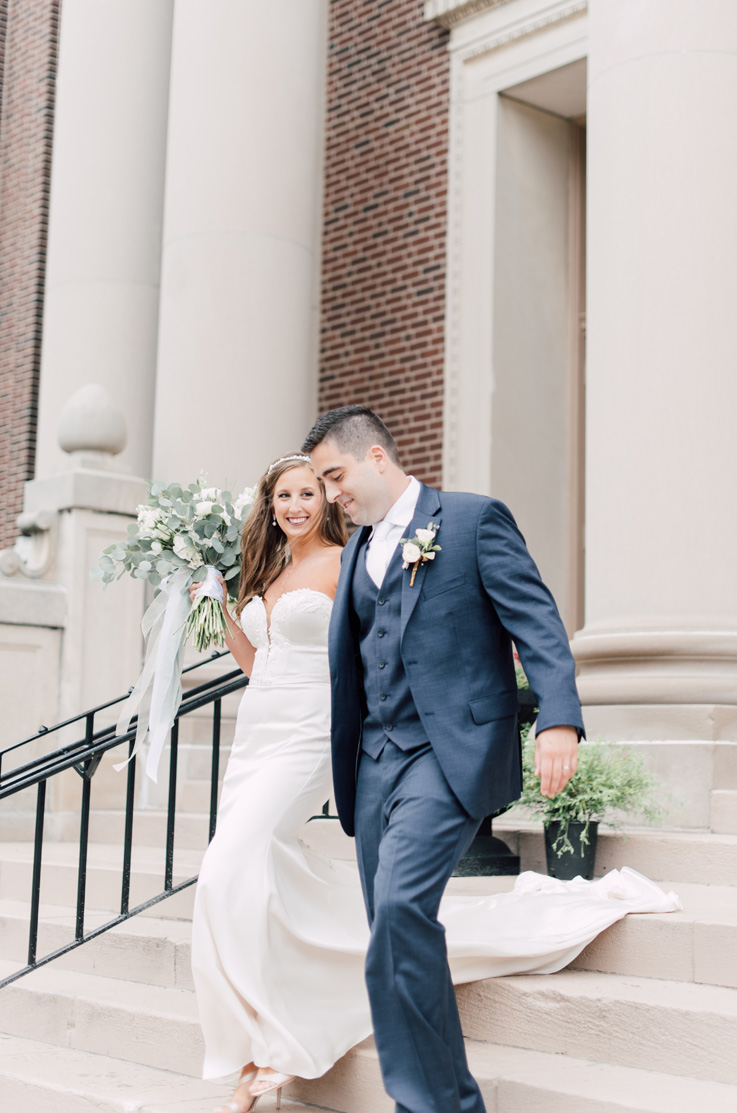 A bride in a plunging neckline wedding dress, holding a bouquet, and a groom in a suit, walk down a cement staircase outside of a brick building.