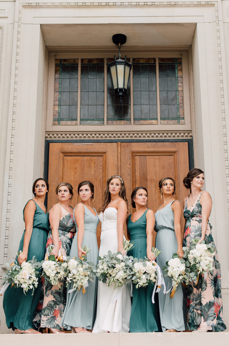 A bride in a plunging neckline wedding dress poses with six bridesmaids in front of the outside of a building with large wooden double doors, all of them are holding bouquets.