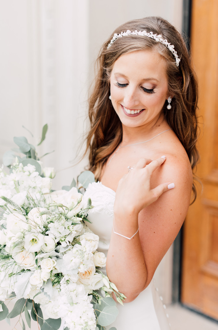 A bride in a strapless wedding dress and a jeweled headband holds a bouquet while posing outside of a building.