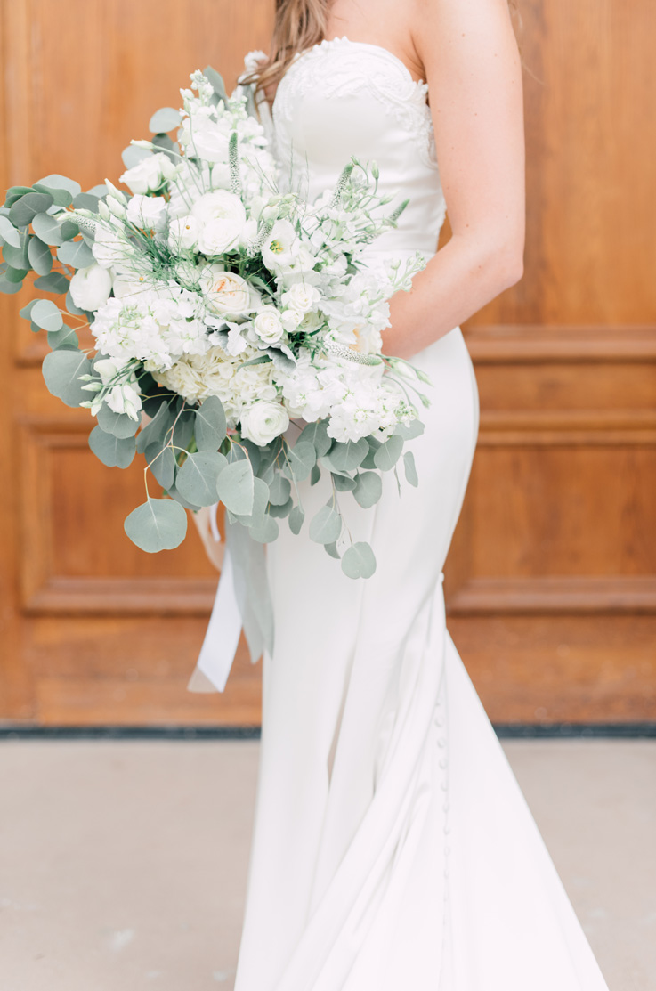 A bride in a strapless fitted wedding dress holds a bouquet and poses in front of a building with wooden doors.