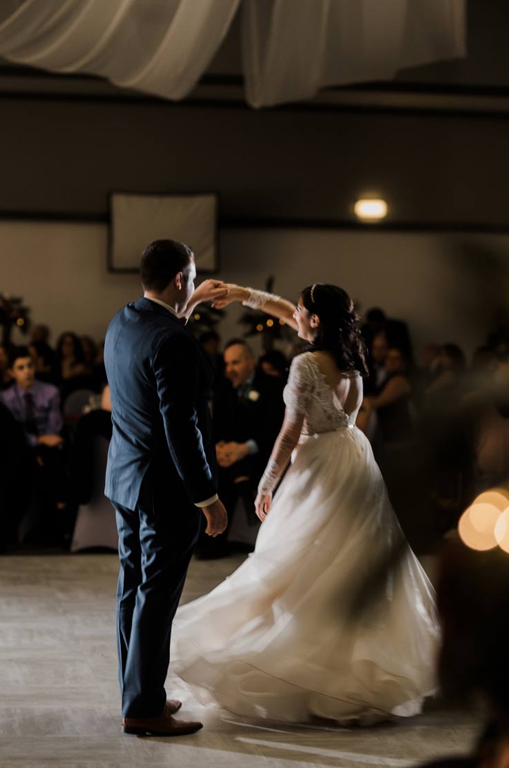 A groom in a suit twirls a bride in a long sleeve ball gown wedding dress on a dance floor inside an event hall, with wedding guests watching in the background.
