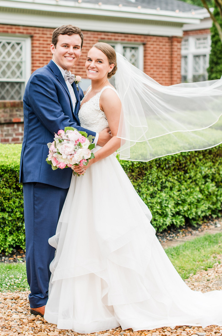 A bride in a tulle skirt wedding dress and veil, holding a bouquet, stands with a groom in a suit outside, with a hedge and a house in the background.