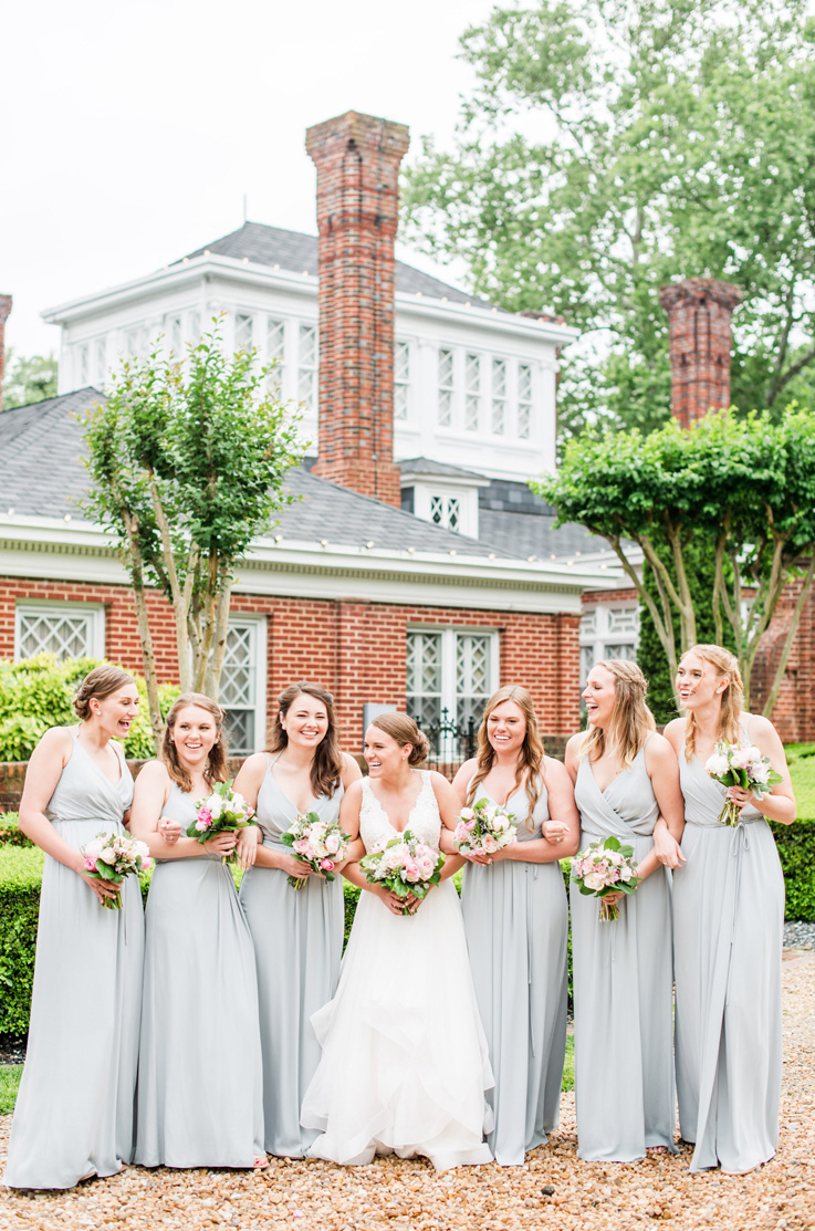 A bride in a V-neck wedding dress stands with six bridesmaids outside of a house, all holding bouquets and laughing with each other.