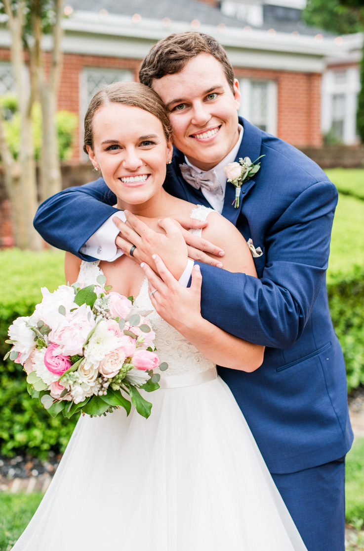 A groom in a suit wraps his arms around the shoulders of a bride in a tulle skirt wedding dress, both of them smiling, with a house and a yard in the background.