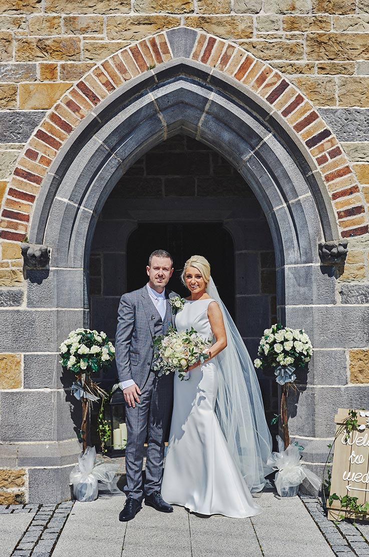 A bride in a sleeveless wedding dress and veil holds a bouquet and stands with a groom in front of a stone archway.
