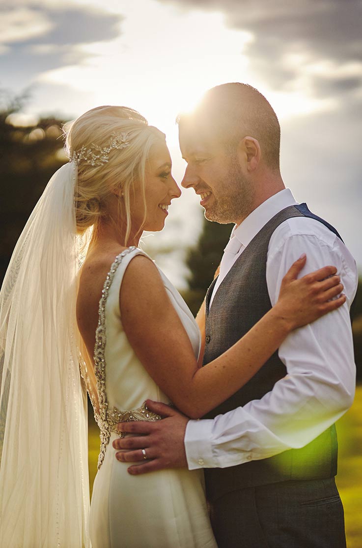 A bride in a sleeveless embellished wedding dress and veil, and a groom in a suit jacked hold each other and smile, while the sun shines behind them.