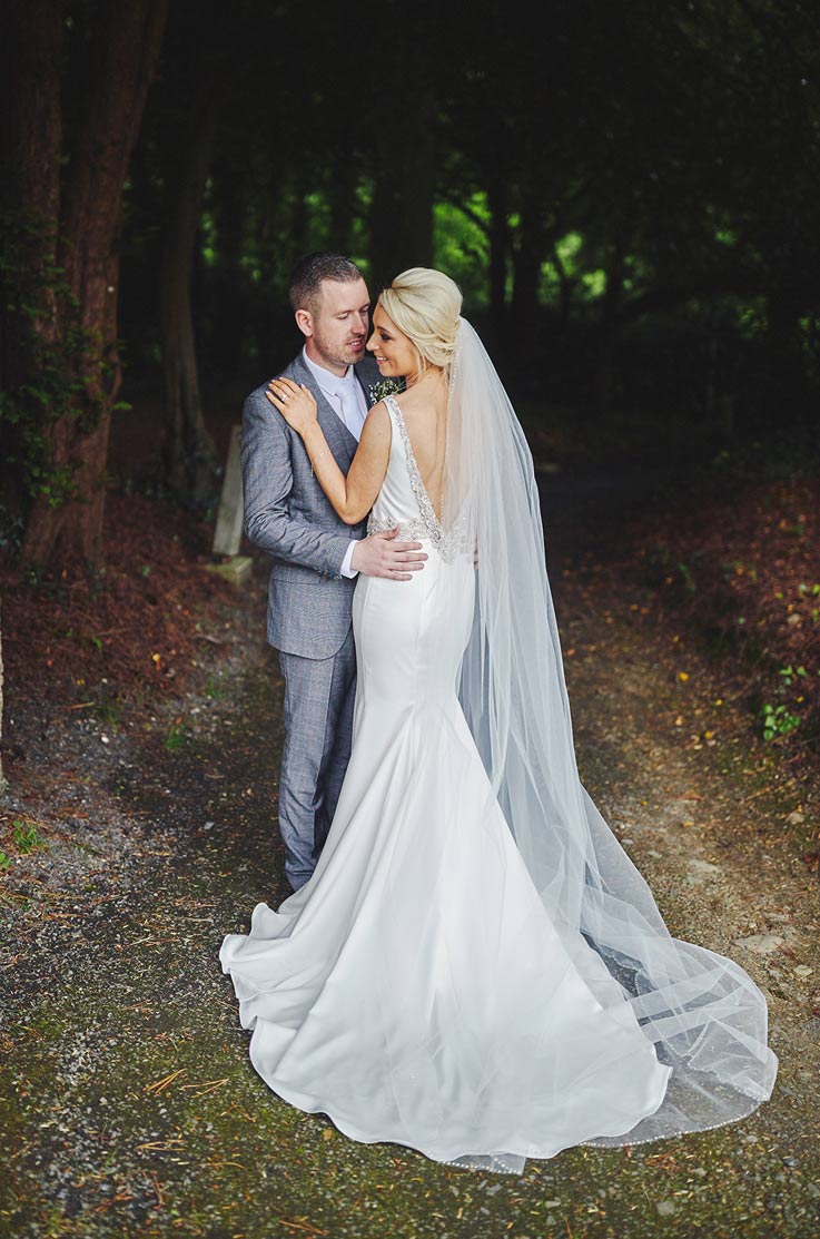 A bride in a sleeveless embellished wedding dress and veil and a groom in a suit stand on a path in a forest and hold each other.