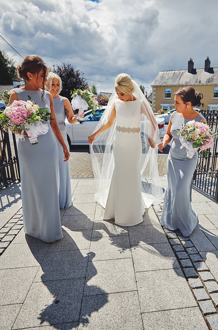 A bride in a sleeveless embellished wedding dress and veil, and three bridesmaids walk down a stone paver path with cars and houses behind them.