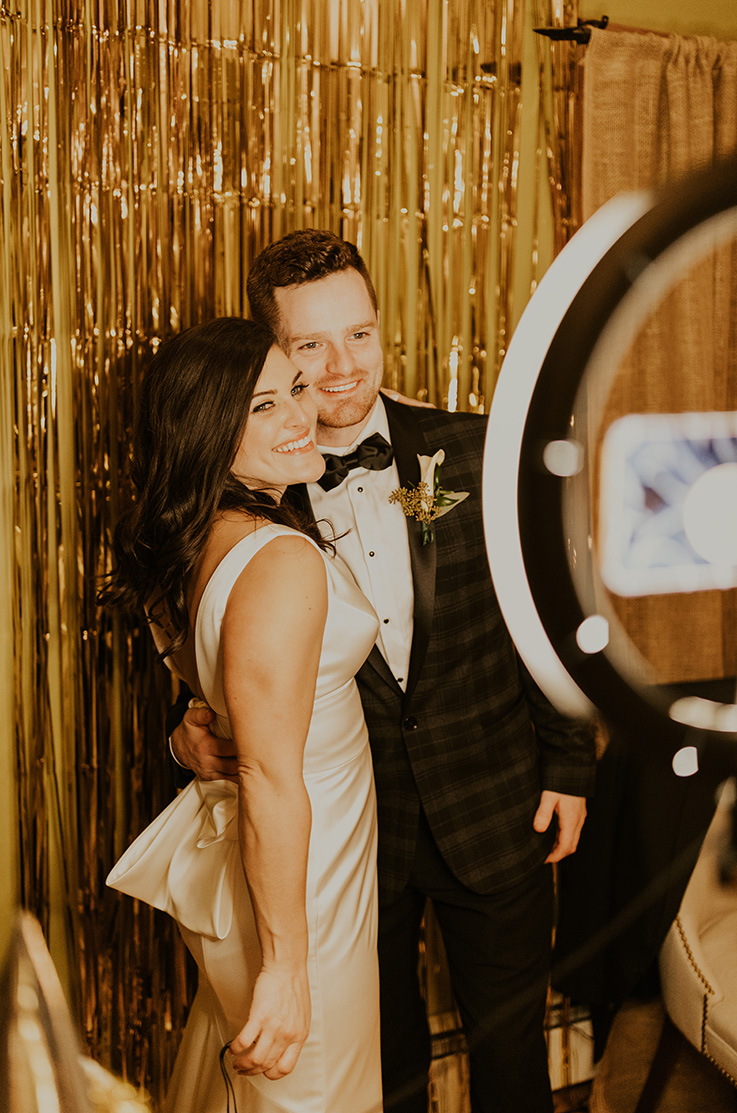 A bride in a sleeveless wedding dress and a groom in a suit pose in front of a glittery backdrop.