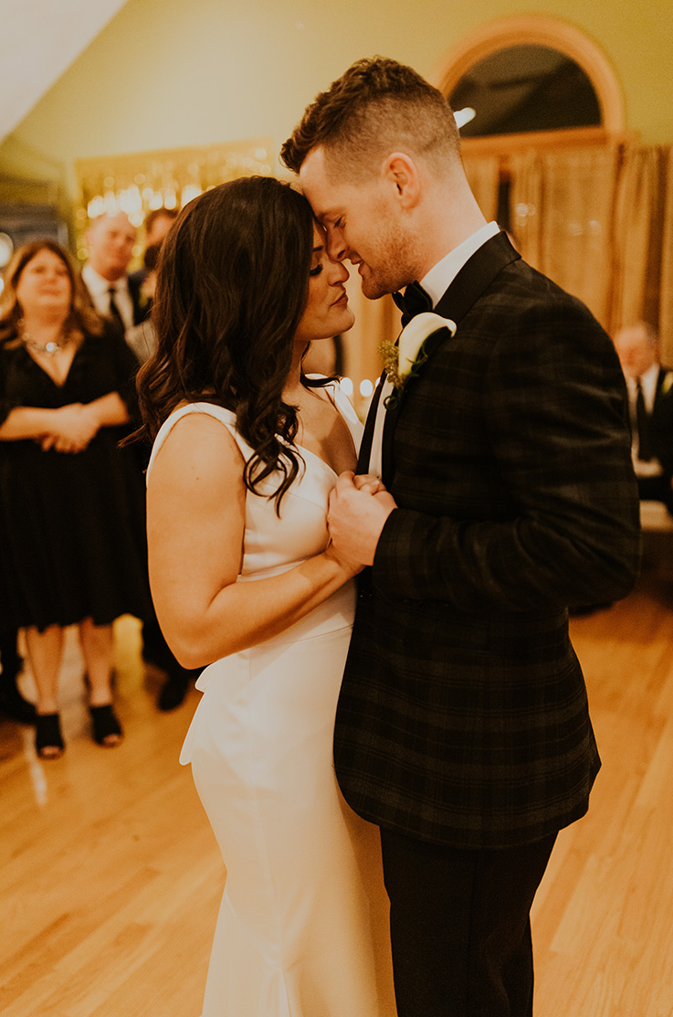 A bride in a sleeveless wedding dress and a groom in a suit dance while wedding guests watch in the background.
