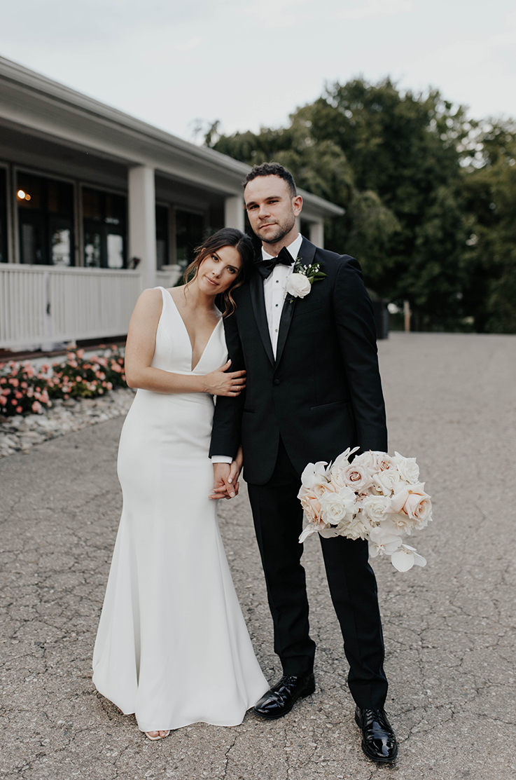 A bride in a sleeveless wedding dress rests her head on the shoulder of a groom in a suit as they stand outside of a building with a covered patio.