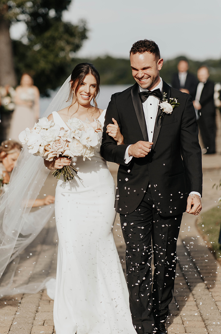 A bride in a fitted wedding dress and veil holding a bouquet while walking down a path with a groom in a suit as wedding guests watch and toss confetti.