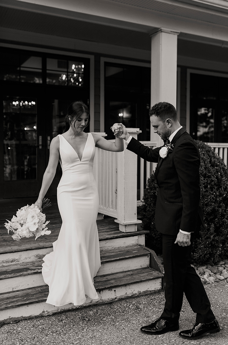 A groom in a suit helps a bride in a V-neck wedding dress down the stairs of a covered patio.