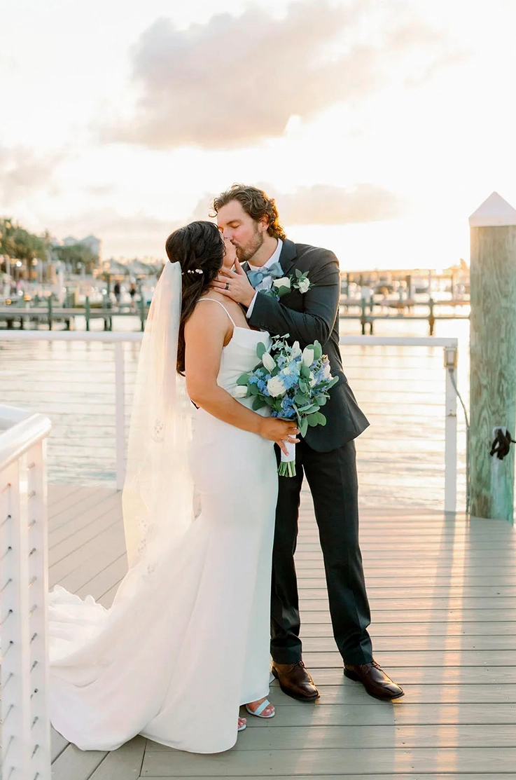 Groom embraces bride in a crepe fit and flare gown with thin straps and a veil, and they kiss on a dock as the sun sets.