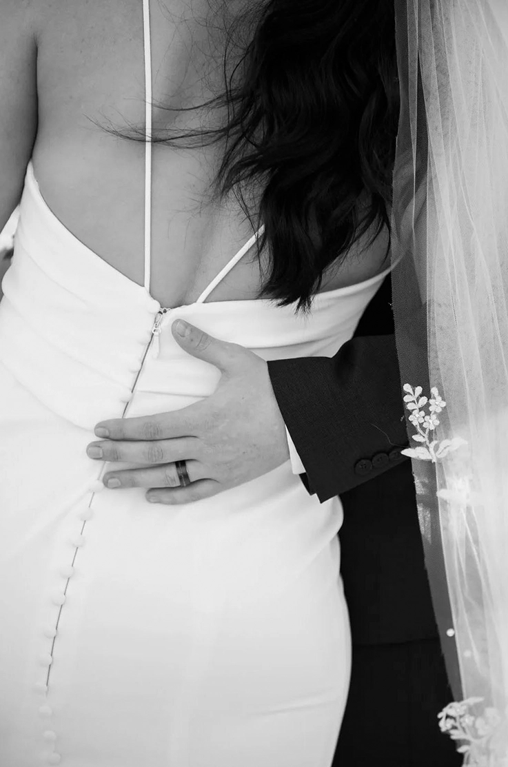 Close-up of groom embracing bride in a buttoned fitted crepe gown with a low back, thin straps and a lace trim veil. 